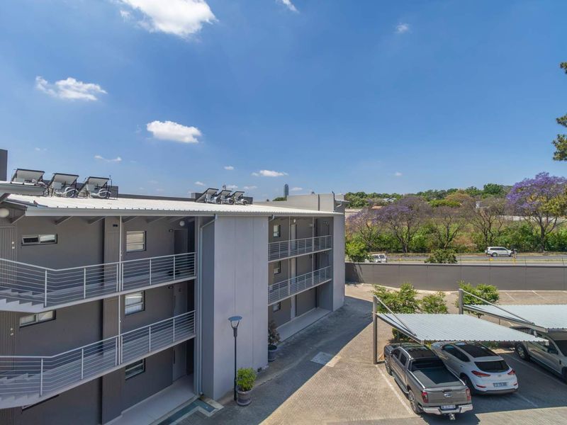 Exterior view of a modern multi-story apartment complex featuring steel balustrades, solar water heating panels, and a paved parking area with carports.