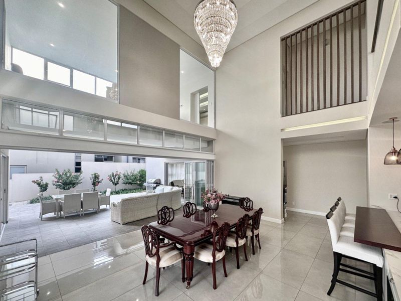 Spacious open-plan dining area featuring double-volume ceilings, a grand crystal chandelier, and seamless flow to the outdoor patio through large sliding glass doors.