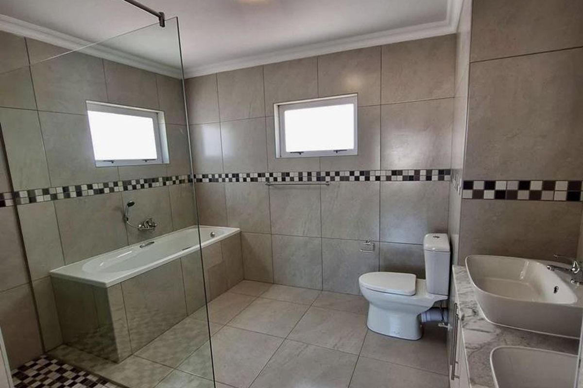 A modern bathroom featuring a glass-enclosed bathtub, dual vanity sinks, and a toilet, finished with neutral wall tiles and decorative mosaic accents.