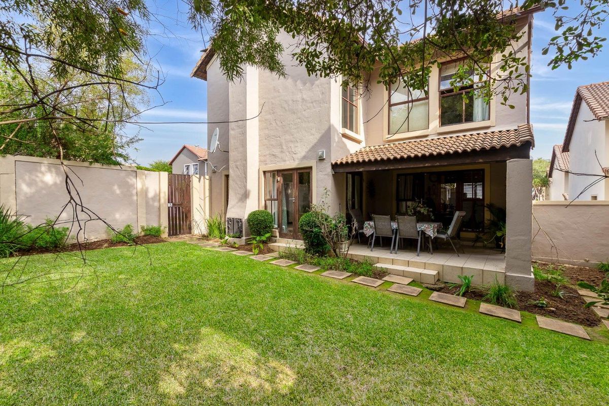 A rear view of the property featuring a covered patio area, tiled flooring, and a well-maintained garden with a lawn and perimeter wall.
