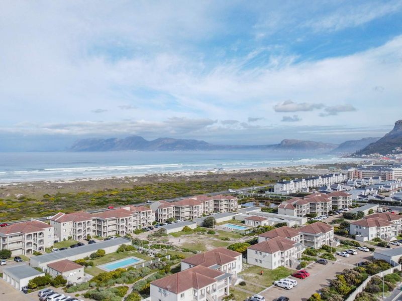 Aerial view of the apartment complex featuring multiple buildings, swimming pools, and paved parking areas, set against a coastal mountain and beach backdrop.