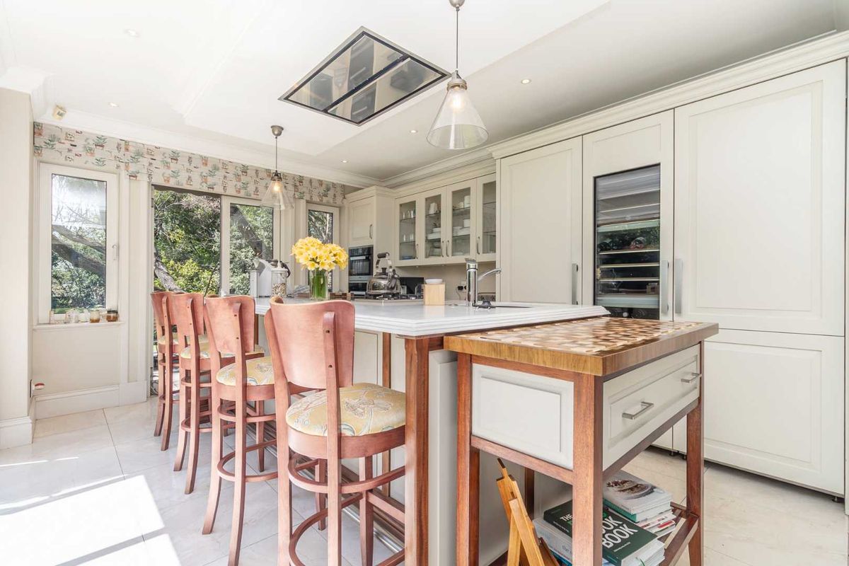 A bright, modern kitchen featuring a large island with stone countertops, integrated wine cooler, and high-end ceiling-mounted extractor fan, opening to a garden view.