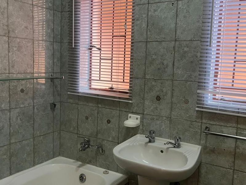 A functional bathroom featuring a bathtub, pedestal basin, and full-height ceramic wall tiling with dual windows and venetian blinds.