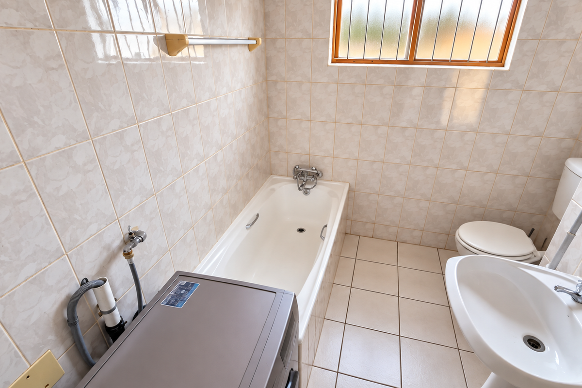A functional bathroom featuring a bathtub, pedestal basin, and toilet, finished with neutral wall and floor tiles and a window with security bars.