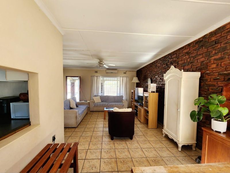 Spacious living room featuring an exposed brick feature wall, tiled flooring, ceiling fan, and air conditioning, with a convenient pass-through window to the kitchen.