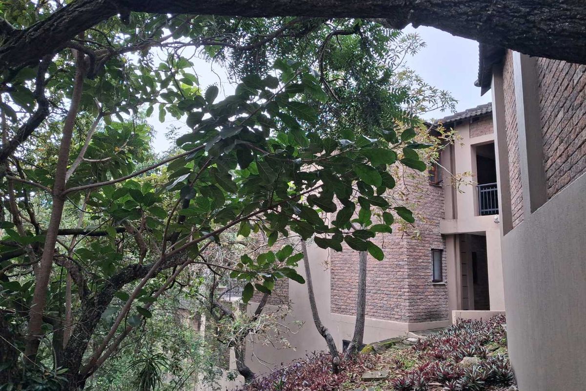 A view of the apartment building exterior featuring brick facade detailing, balconies, and surrounding mature trees and landscaped garden areas.