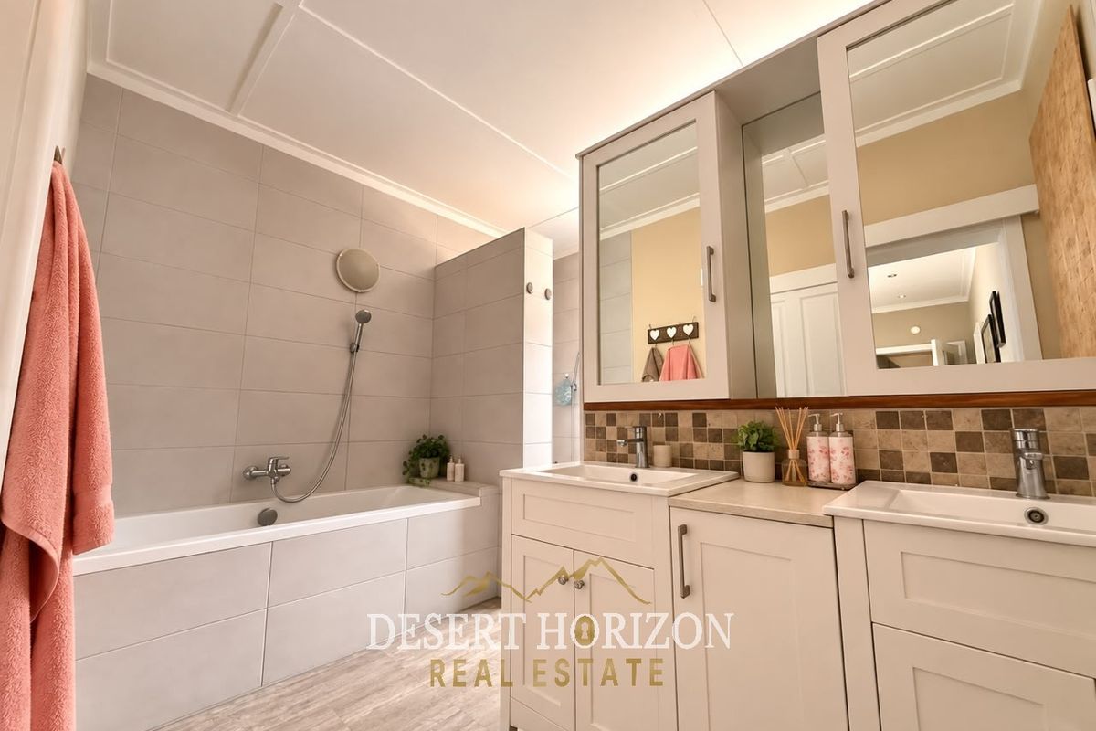 Modern bathroom featuring a double vanity with mirrored cabinets, a tiled bathtub with shower attachment, and neutral wall tiling.