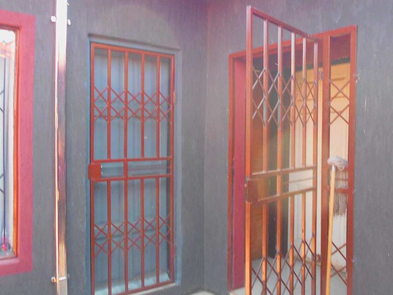 A rear exterior area featuring tiled flooring, security gates on doorways, and a downpipe against a dark-painted wall.