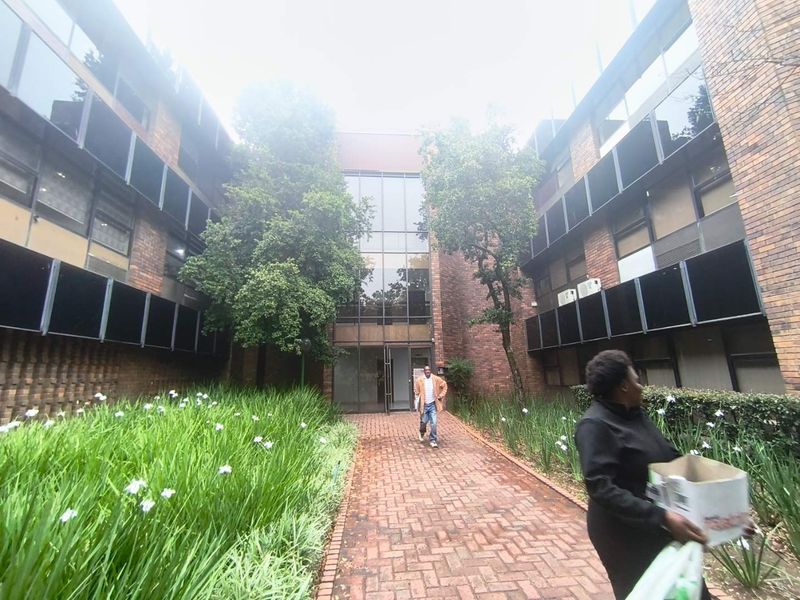 Front entrance view of a multi-story office building featuring a brick facade, glass curtain wall, and a paved walkway through a landscaped garden.