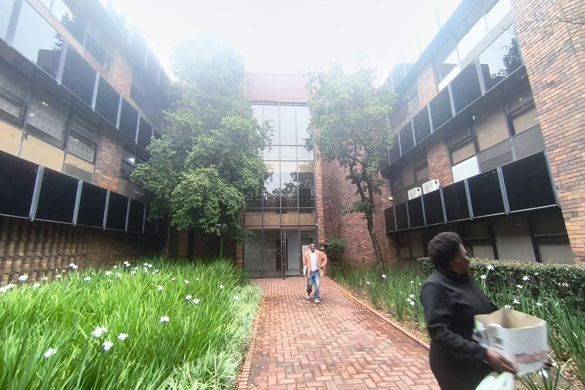 Front entrance view of a multi-story office building featuring a brick facade, glass curtain wall, and a paved walkway through a landscaped garden.