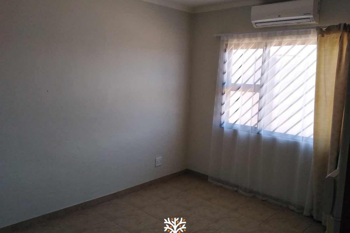 A clean, empty bedroom featuring tiled flooring, a wall-mounted air conditioning unit, and a window with fitted security bars.