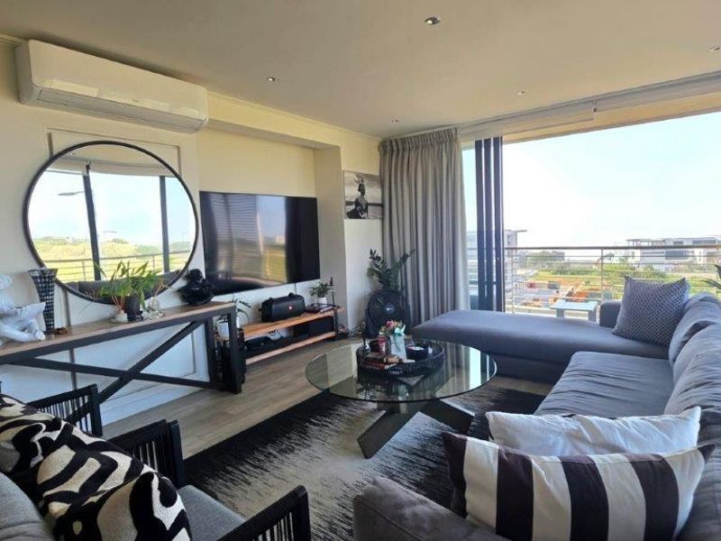 A bright and modern living room featuring laminate flooring, recessed lighting, a wall-mounted air conditioner, and large sliding doors leading to a balcony.