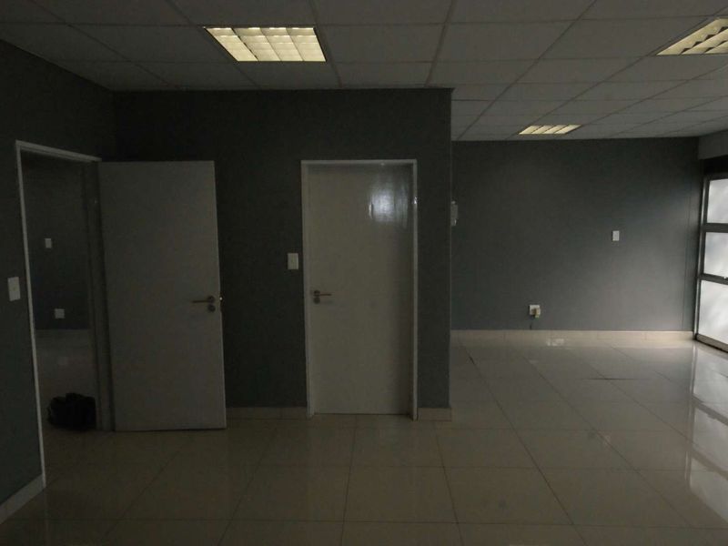 An empty office space featuring tiled flooring, recessed lighting in a suspended ceiling, and neutral grey walls, providing a clean and professional work environment.