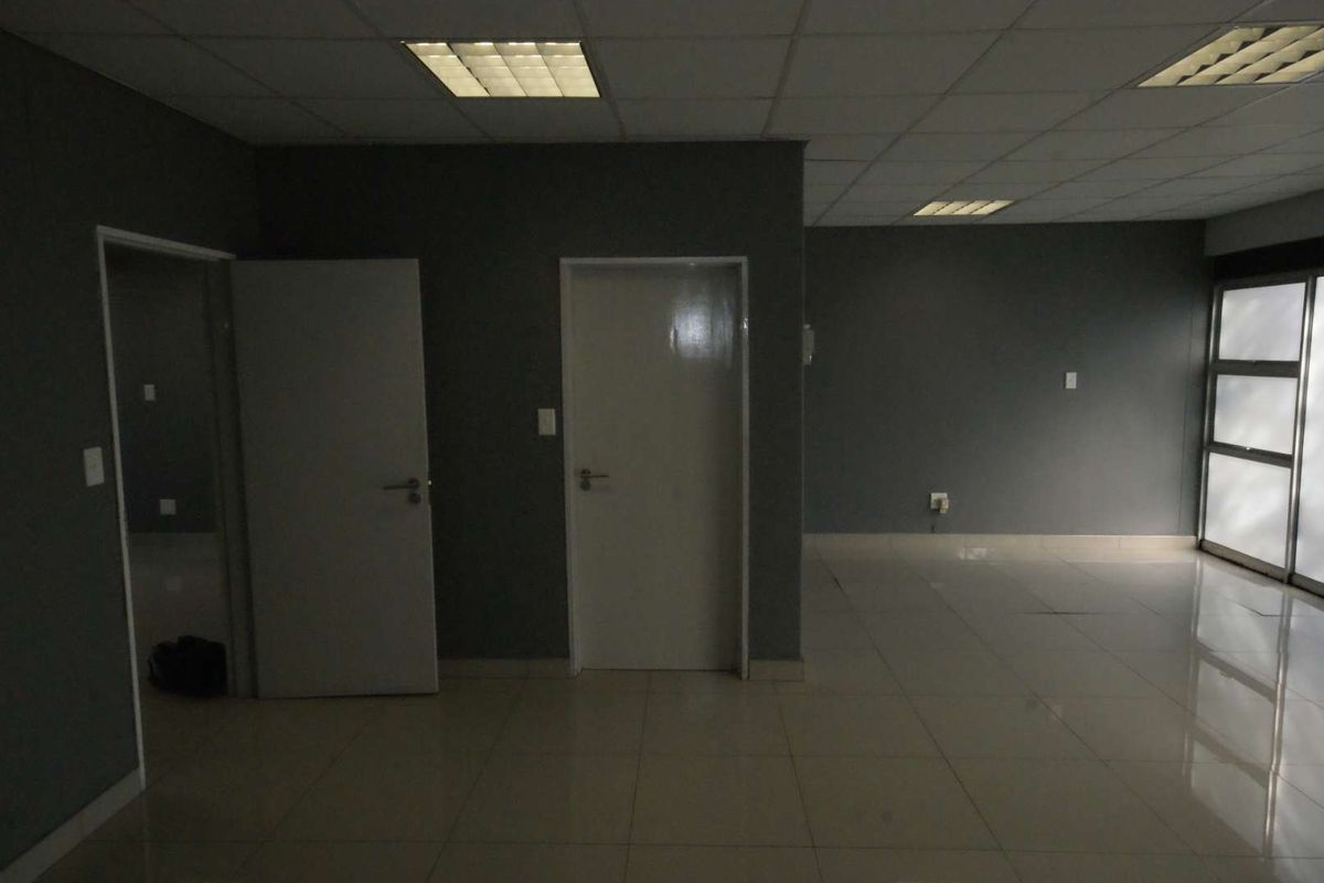 An empty office space featuring tiled flooring, recessed lighting in a suspended ceiling, and neutral grey walls, providing a clean and professional work environment.