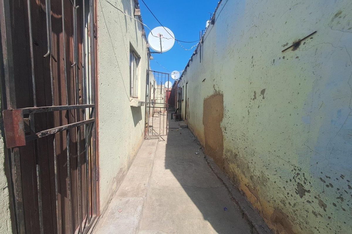 A narrow exterior side passage between two structures featuring concrete flooring, security gates, and visible wear to the exterior wall finishes.