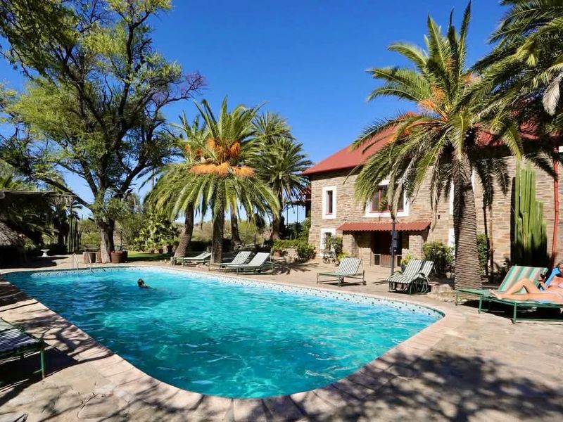 A well-maintained outdoor pool area featuring stone paving, mature palm trees, and a stone-clad farmhouse building in the background.