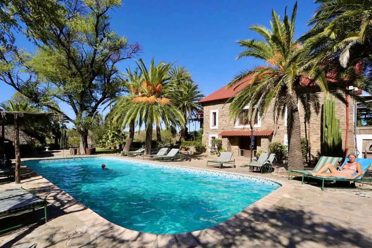 A well-maintained outdoor pool area featuring stone paving, mature palm trees, and a stone-clad farmhouse building in the background.