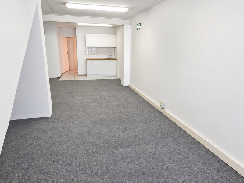 An open-plan industrial office space featuring a small kitchenette area with built-in cabinetry and sink, finished with neutral carpeting and white walls.