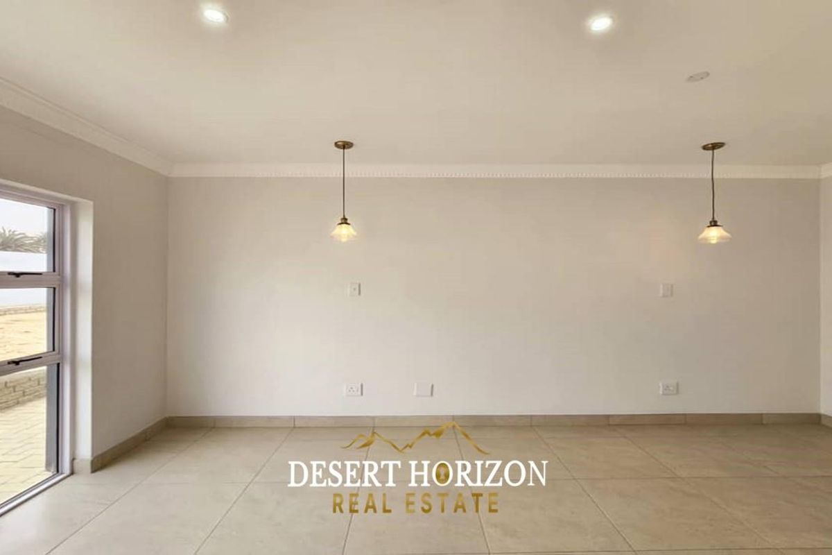 A bright, open-plan living area featuring neutral tiled flooring, recessed ceiling lighting, and two decorative pendant lights, with access to an outdoor patio.