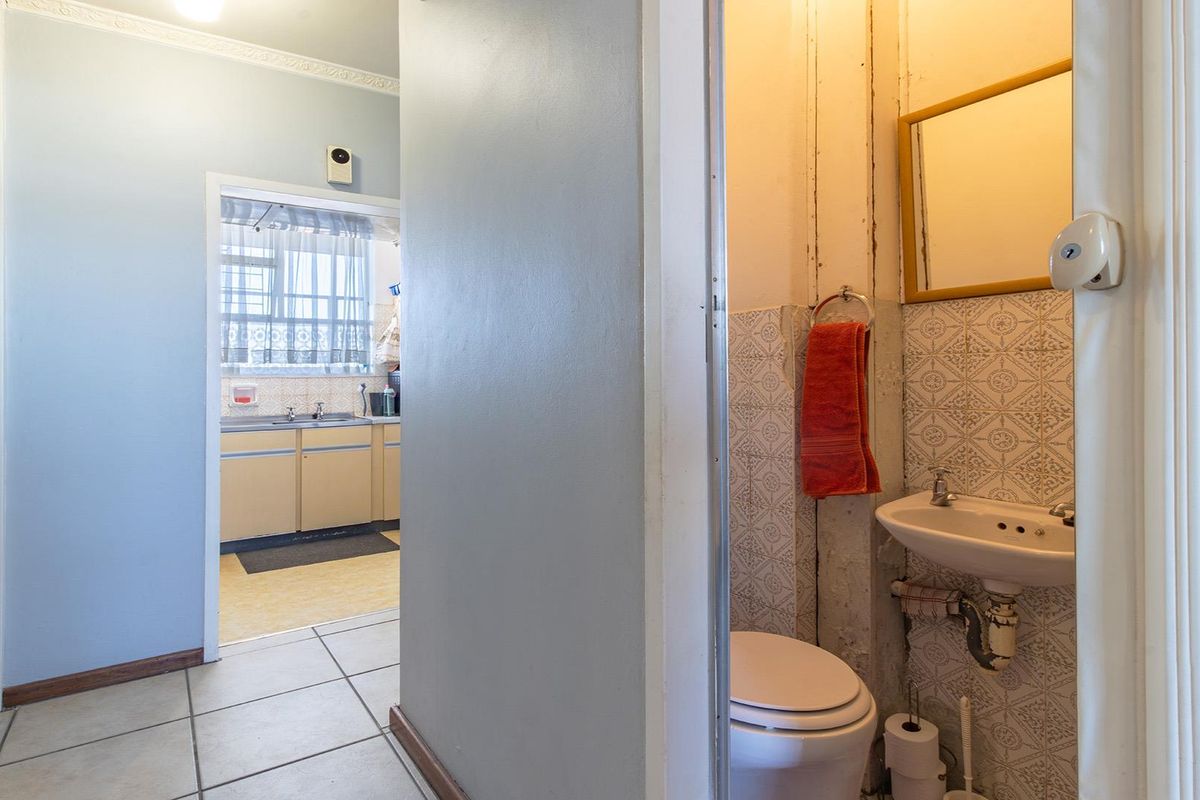 A compact bathroom featuring a pedestal basin and toilet, with visible wear to wall finishes and plumbing, adjacent to a kitchen area.