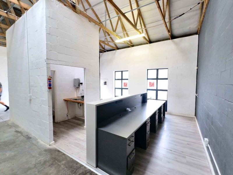 Open-plan industrial office space featuring exposed timber roof trusses, large steel-framed windows, and a built-in workstation area with adjacent utility nook.
