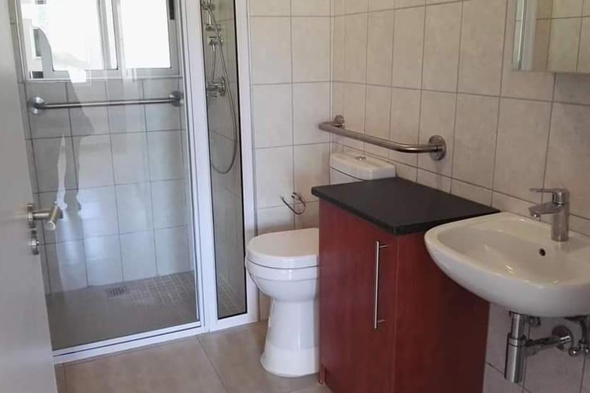 A functional bathroom featuring a glass-enclosed shower, wall-mounted basin, toilet, and safety grab rails, finished with neutral wall tiles.