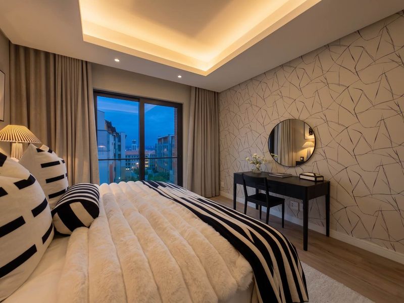 Modern bedroom featuring recessed cove lighting, patterned feature wallpaper, laminate flooring, and a sliding glass door leading to a balcony with urban views.
