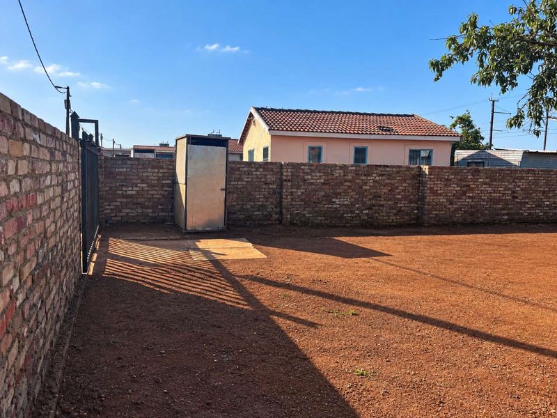 A spacious, enclosed rear yard featuring a brick boundary wall, metal gate, and a small detached metal utility structure on unpaved ground.