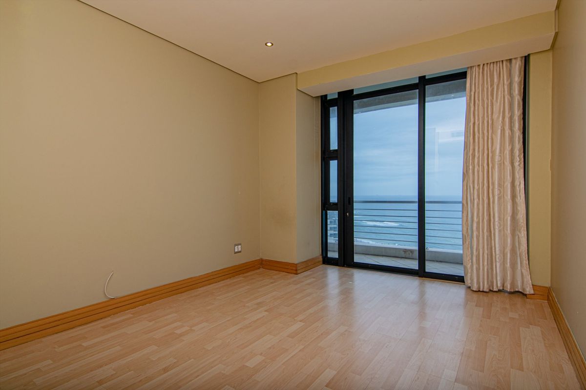A bright, empty bedroom featuring light wood-look flooring, neutral walls, and large sliding glass doors that open onto a balcony with ocean views.