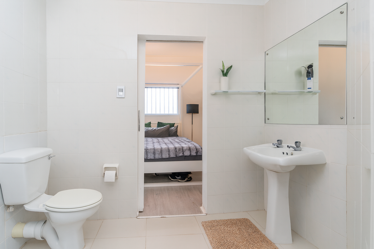 A functional bathroom featuring a pedestal sink, wall-mounted mirror, and toilet, with a direct view into the adjacent bedroom.