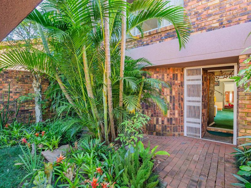 A welcoming front entrance featuring a brick-paved pathway, established garden with palm trees, and a secure metal gate leading into the home.