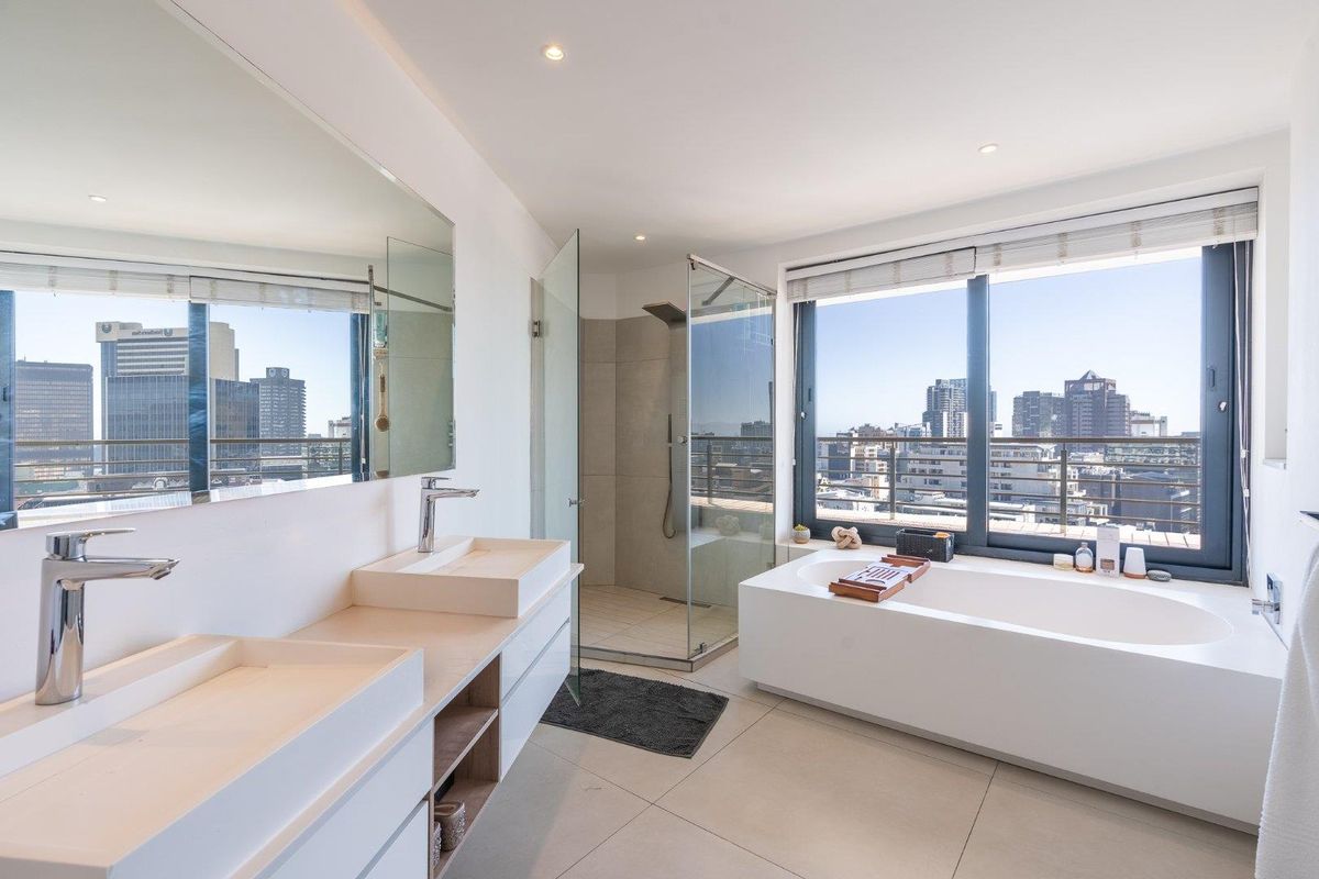 Modern penthouse bathroom featuring a double vanity, walk-in shower, and a freestanding bathtub with expansive city views through large windows.