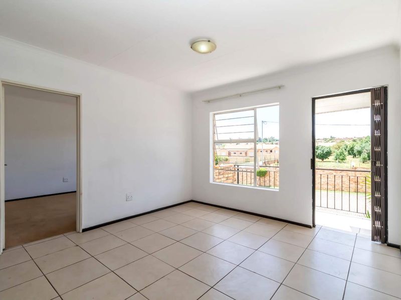 An empty, well-lit living area featuring neutral ceramic floor tiles, a security gate leading to a balcony, and large windows providing natural light.