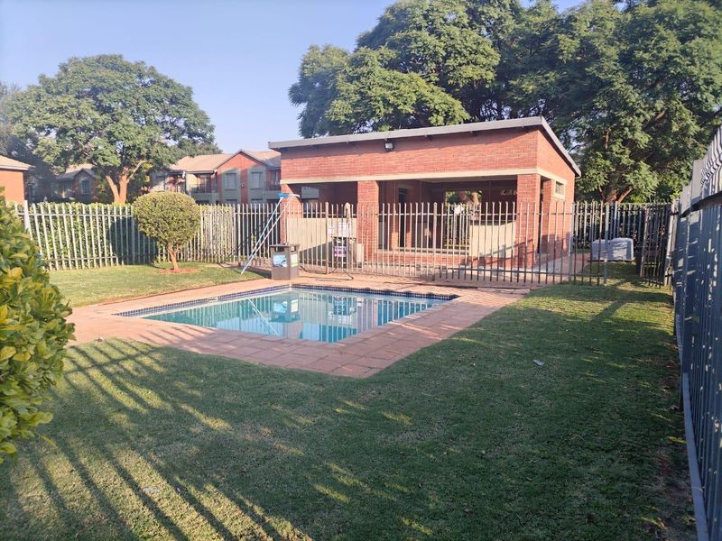 Communal pool area featuring a brick clubhouse, paved surround, and secure metal perimeter fencing within a landscaped garden setting.