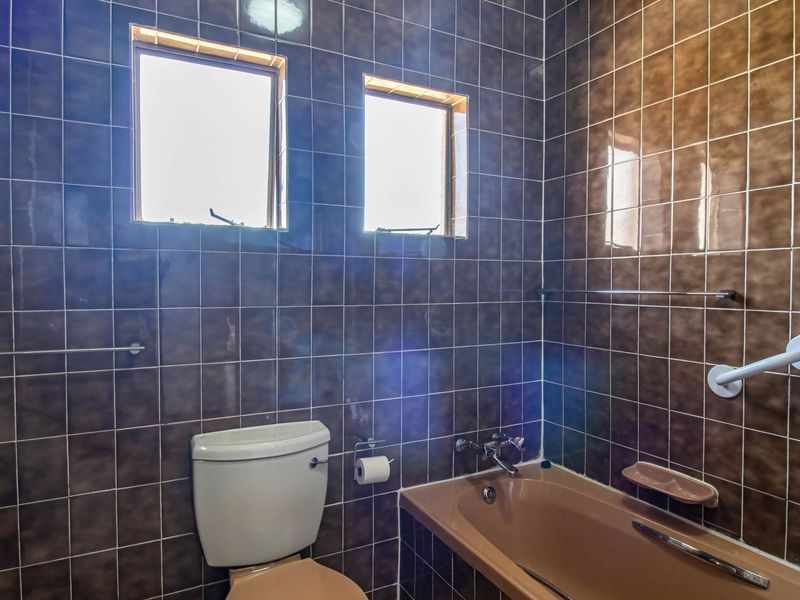 A dated bathroom featuring dark wall tiles, a built-in bathtub, and a toilet, with two small casement windows providing natural light.