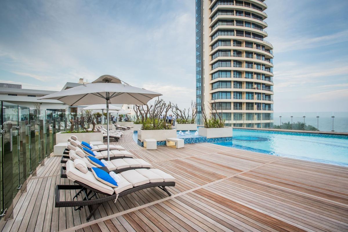 A luxury rooftop swimming pool area featuring timber decking, glass balustrades, and built-in planters with a view of the high-rise apartment building.