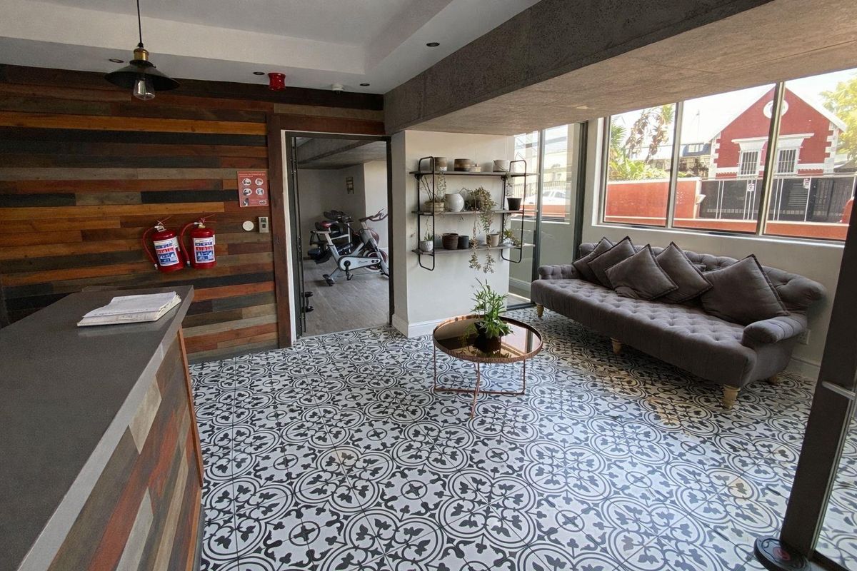 A modern communal lounge area featuring decorative patterned floor tiles, wood-paneled accent wall, and large windows, with an adjacent gym space visible.