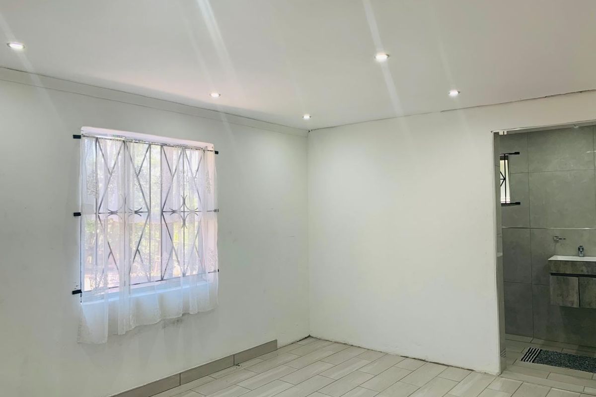 A bright, empty bedroom featuring tiled flooring, recessed downlights, security bars on the window, and direct access to an en-suite bathroom.