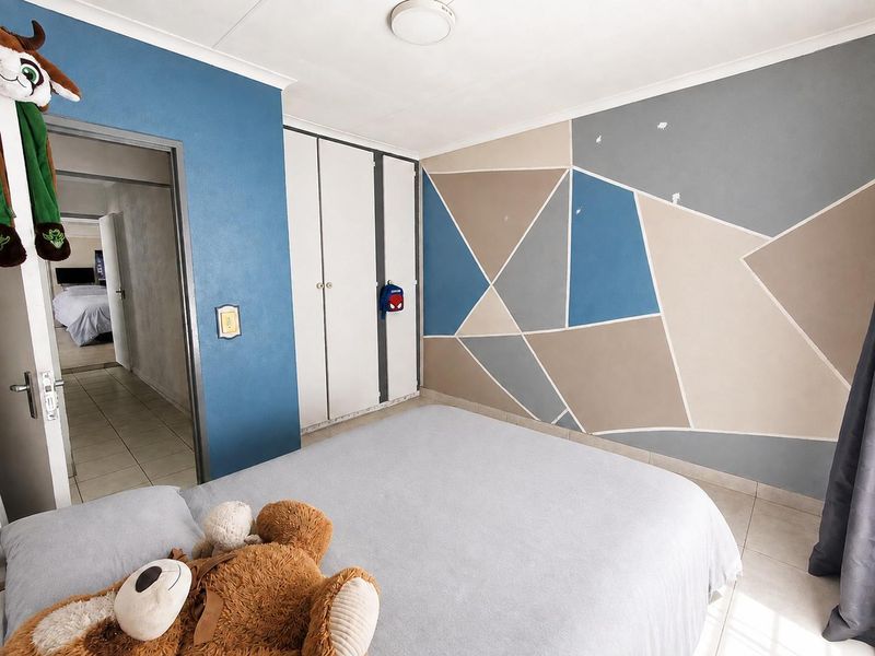 A bedroom featuring built-in cupboards, tiled flooring, and a geometric painted feature wall, offering a functional space with potential for cosmetic updates.