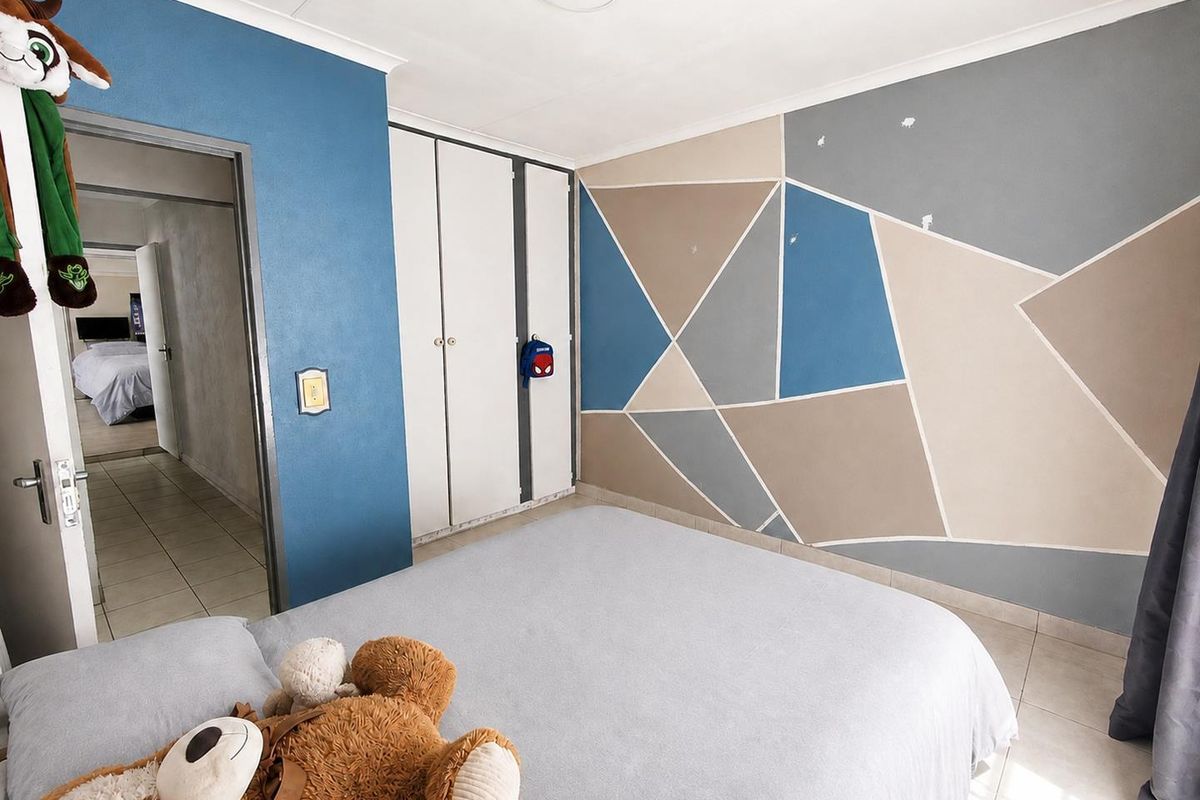A bedroom featuring built-in cupboards, tiled flooring, and a geometric painted feature wall, offering a functional space with potential for cosmetic updates.