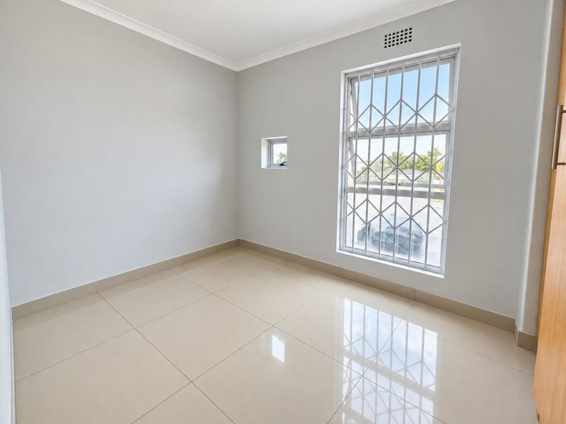 A bright, empty bedroom featuring neutral tiled flooring, built-in cupboard storage, and security bars on the windows.