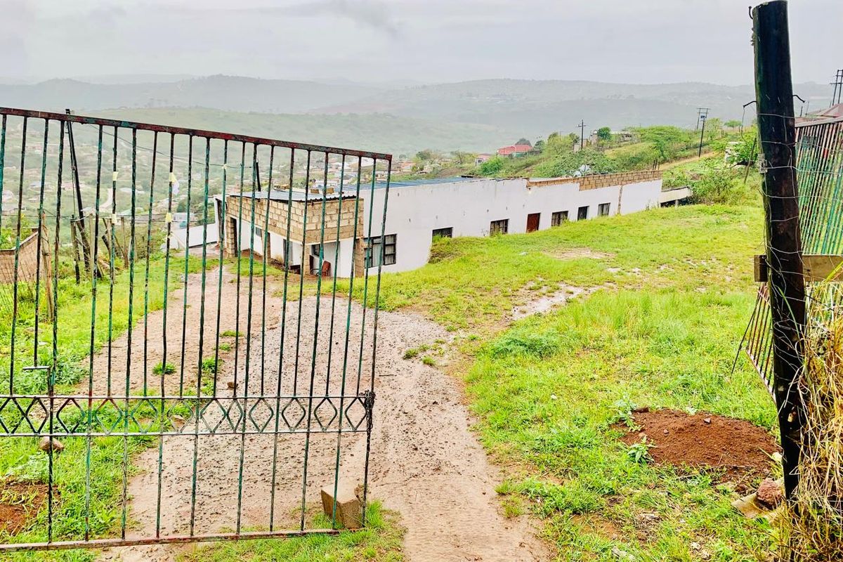 Exterior view of a property featuring a metal security gate, gravel driveway, and unfinished masonry walls set against a hilly landscape.