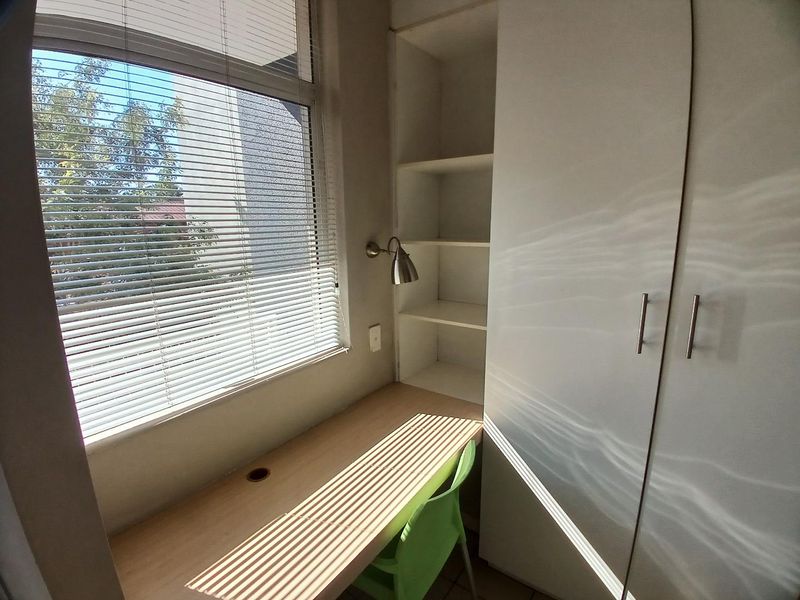 A compact home office featuring a built-in desk, integrated shelving, and a large built-in wardrobe, illuminated by natural light through venetian blinds.