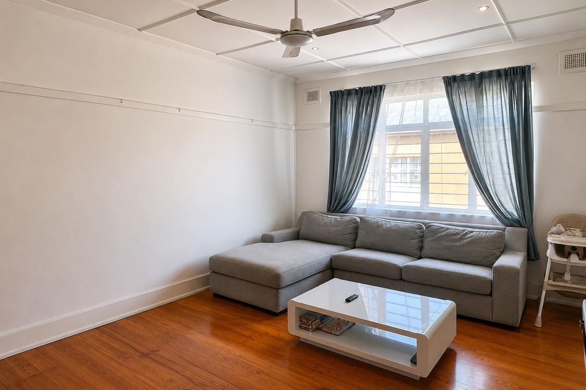 A bright living room featuring polished timber flooring, a classic grid-pattern ceiling with a central fan, and large windows with security bars.