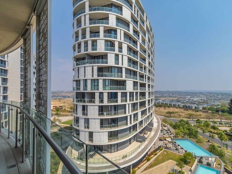 Elevated view from a balcony overlooking a modern high-rise apartment building, swimming pool area, and surrounding landscape.