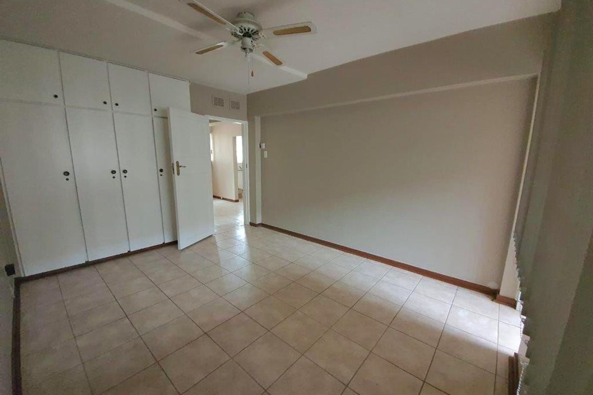 A spacious, empty bedroom featuring neutral tiled flooring, built-in cupboards for storage, and a ceiling fan, offering a blank canvas for personalization.