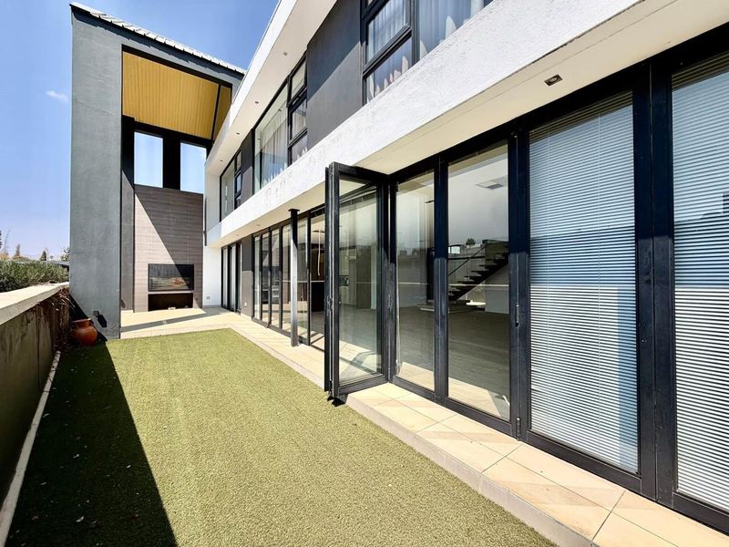 Modern rear exterior featuring a tiled patio, artificial lawn, built-in outdoor fireplace, and expansive bi-fold glass doors leading into the home.