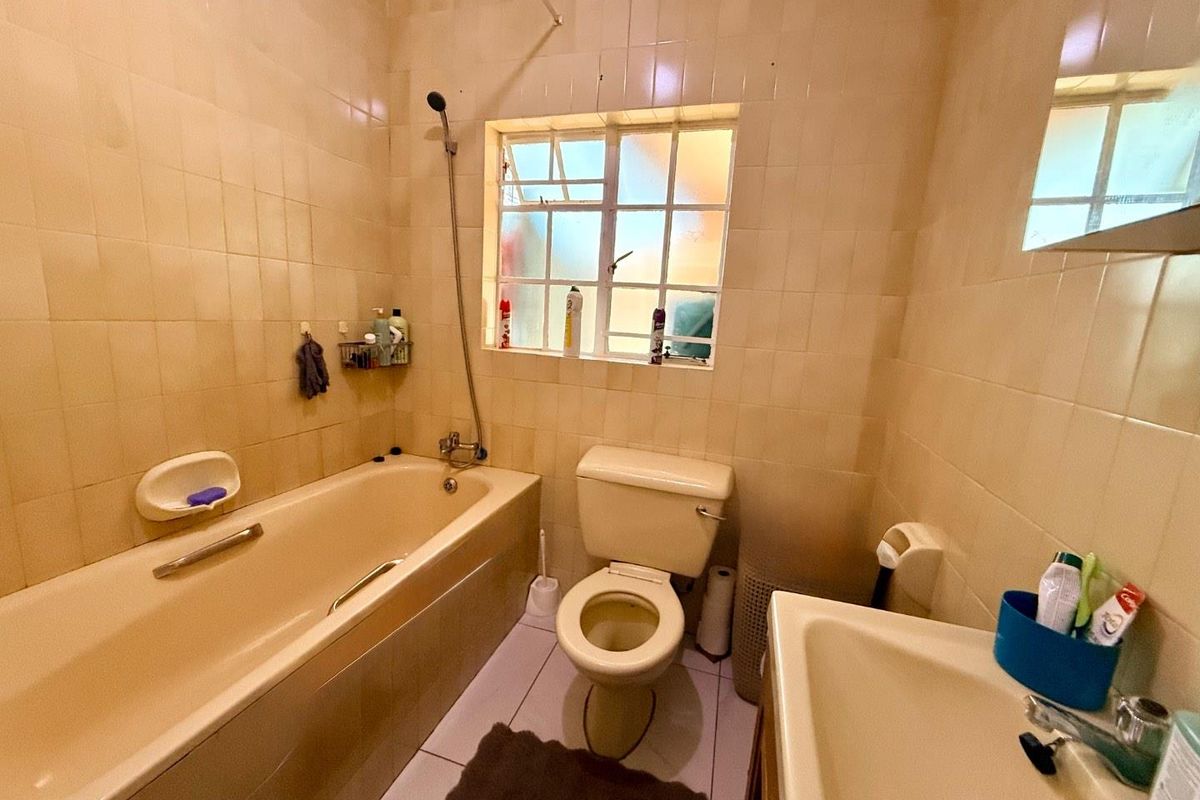 A dated bathroom featuring a built-in bathtub, standard toilet, and vanity basin with full-height wall tiling and a small window for ventilation.