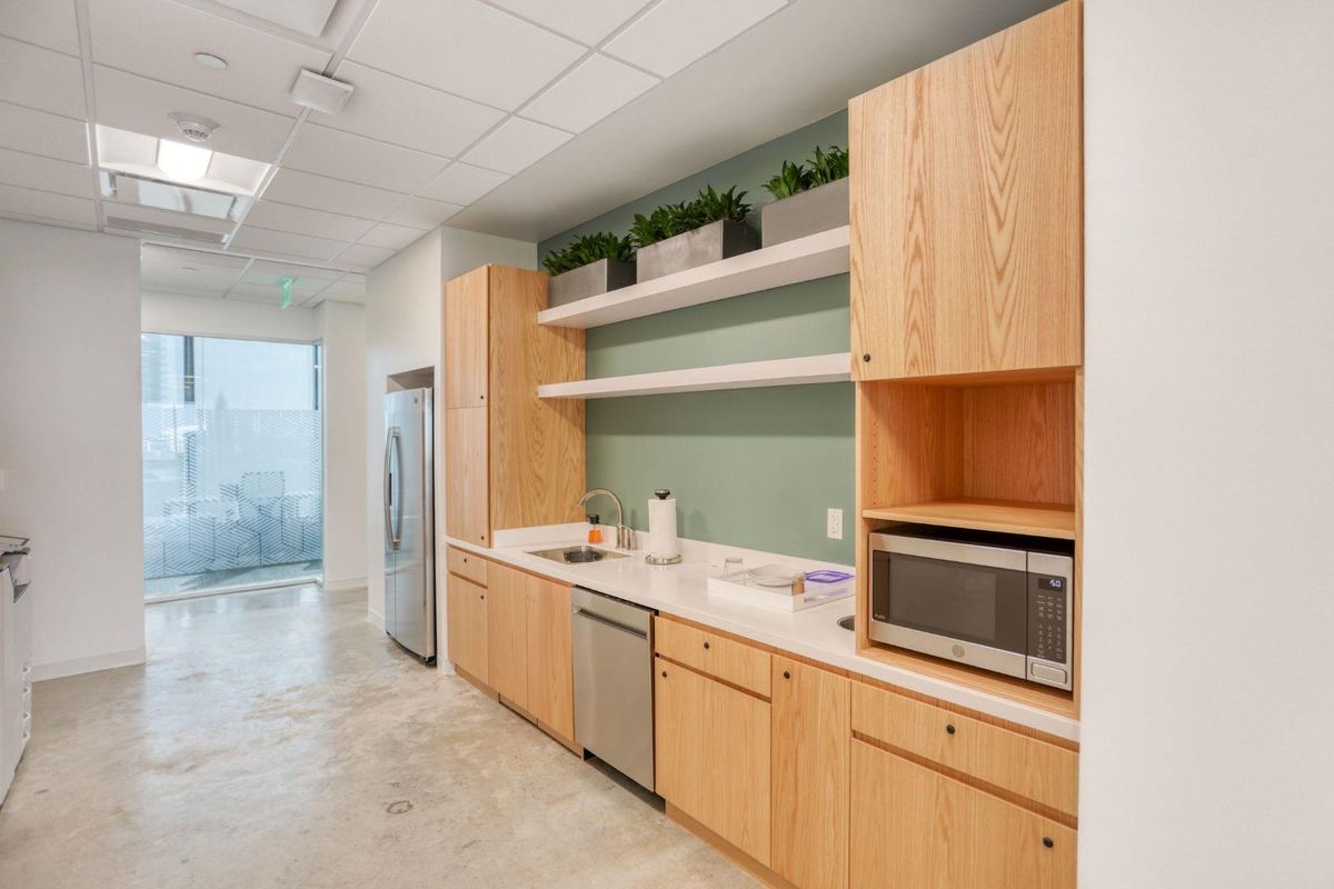 A modern office kitchenette featuring light wood cabinetry, white countertops, a stainless steel dishwasher, and a built-in microwave, set against a soft green accent wall.