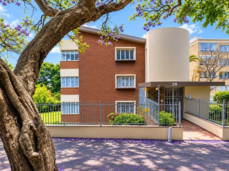 Front exterior view of a multi-story brick apartment building featuring a secure gated entrance and established surrounding trees.